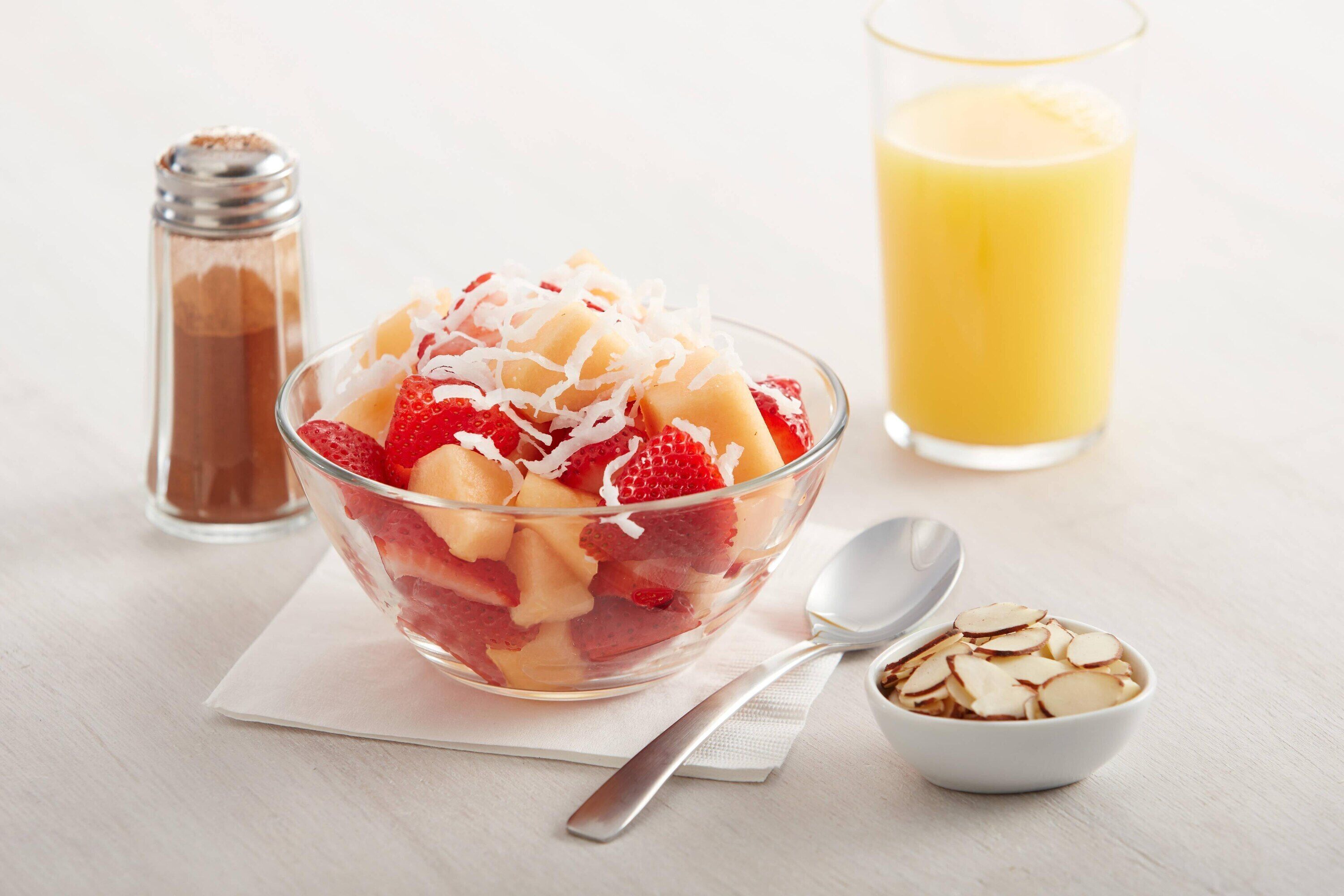 Een fruitsalade met aardbeien en meloen, een glas sinaasappelsap, en een klein schaaltje amandelen op een witte tafel.