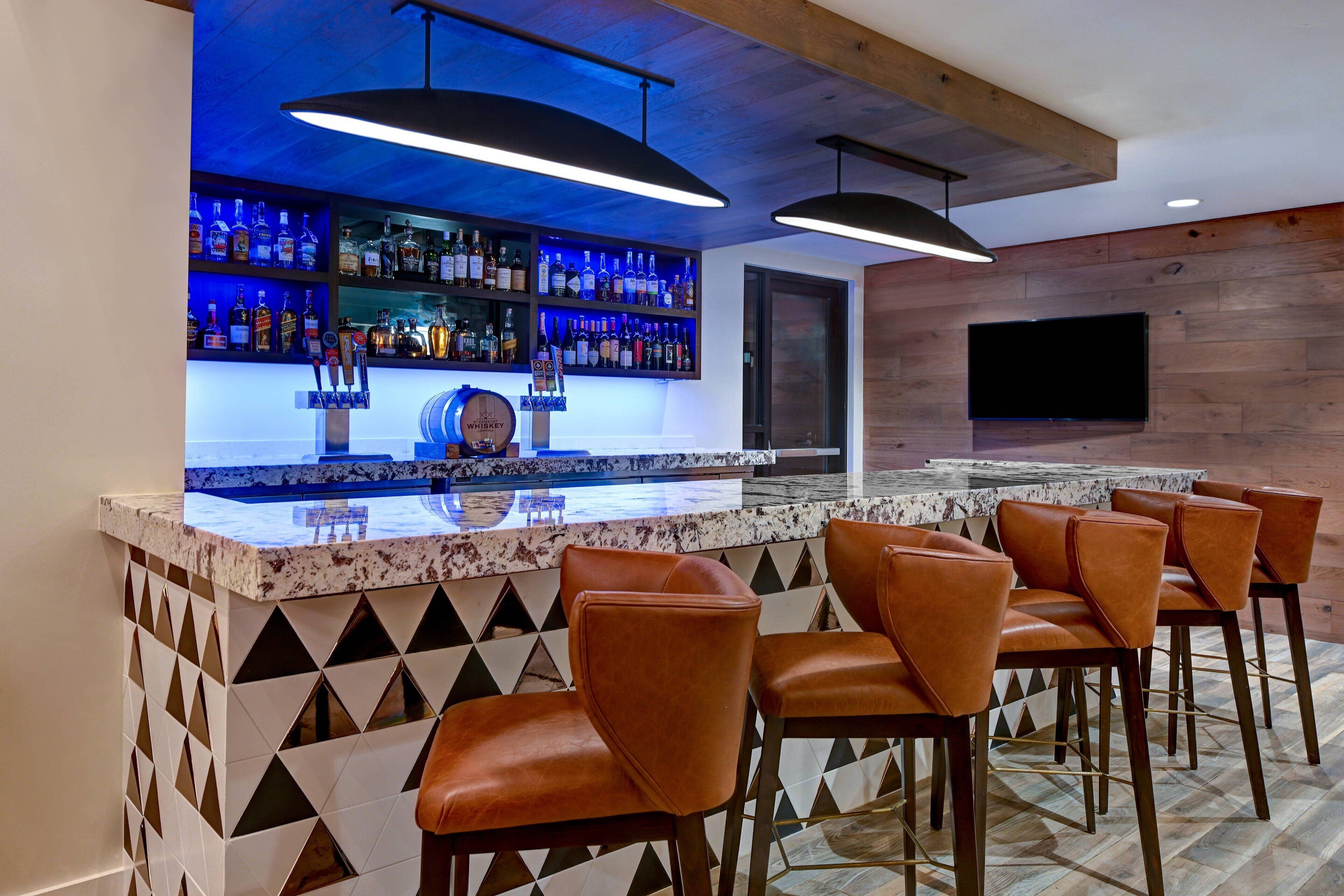 Uma área de bar com bancada de mármore, banquetes de couro e uma prateleira com garrafas de bebidas.