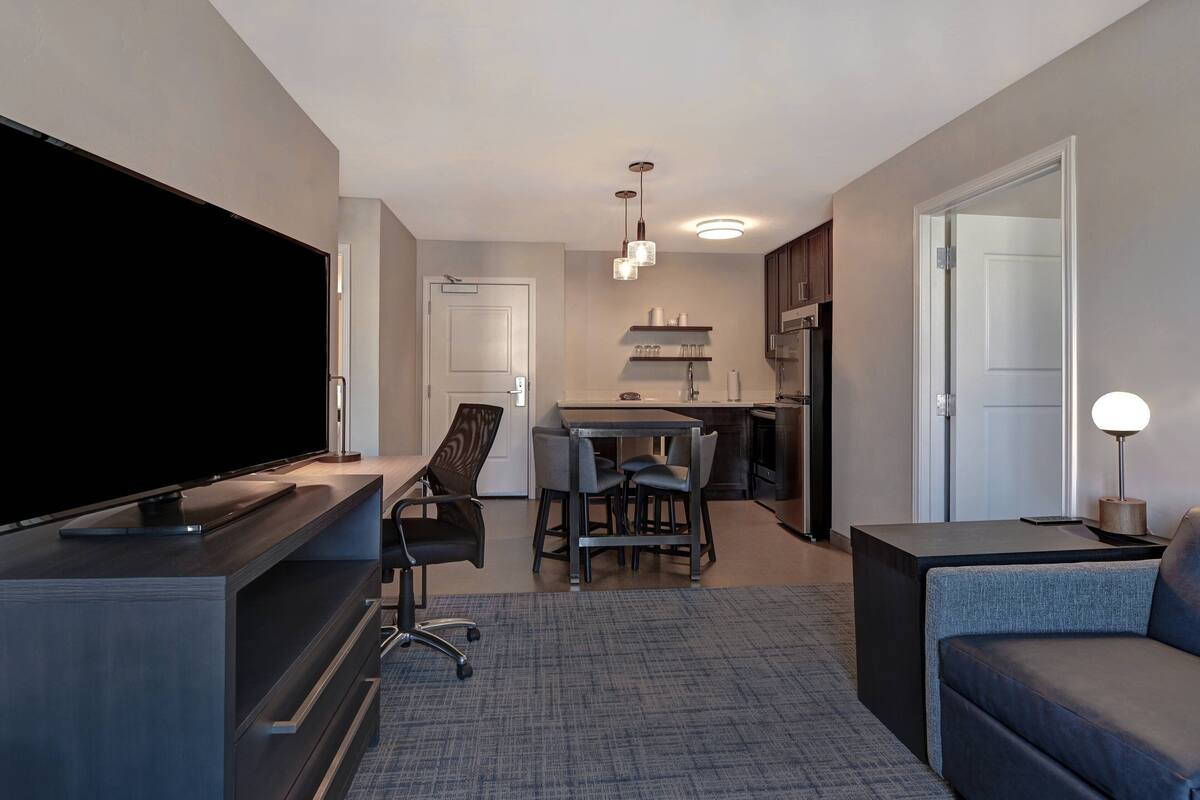 A modern living room with a flat-screen TV, a sofa, a coffee table, and a dining area with chairs and a table.
