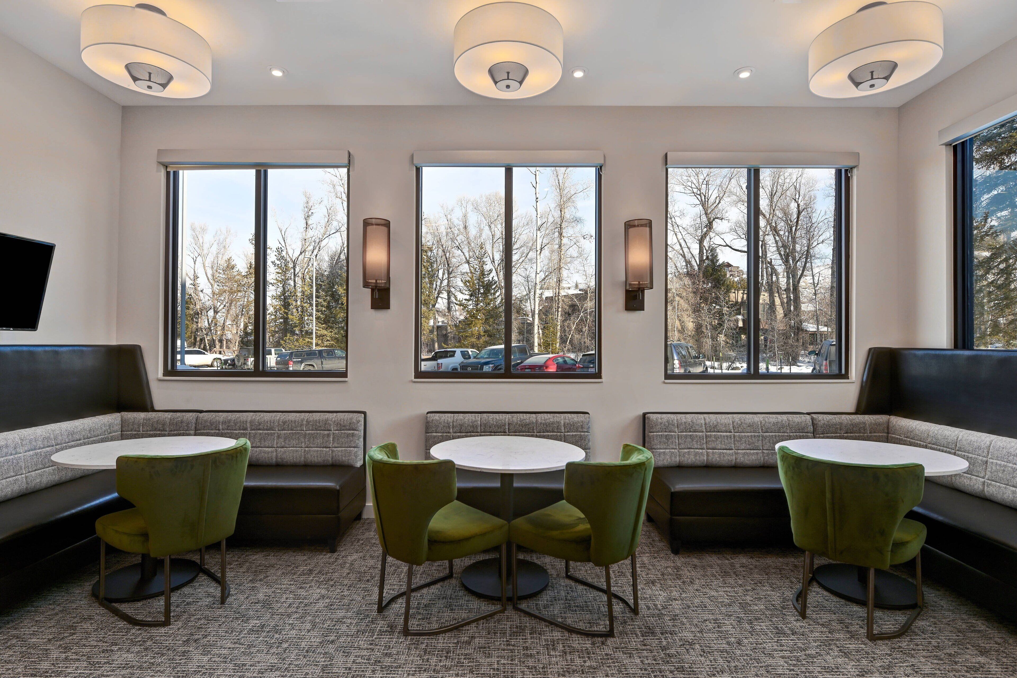 A modern interior with a seating area, a round table, and large windows offering a view of the parking lot and trees.
