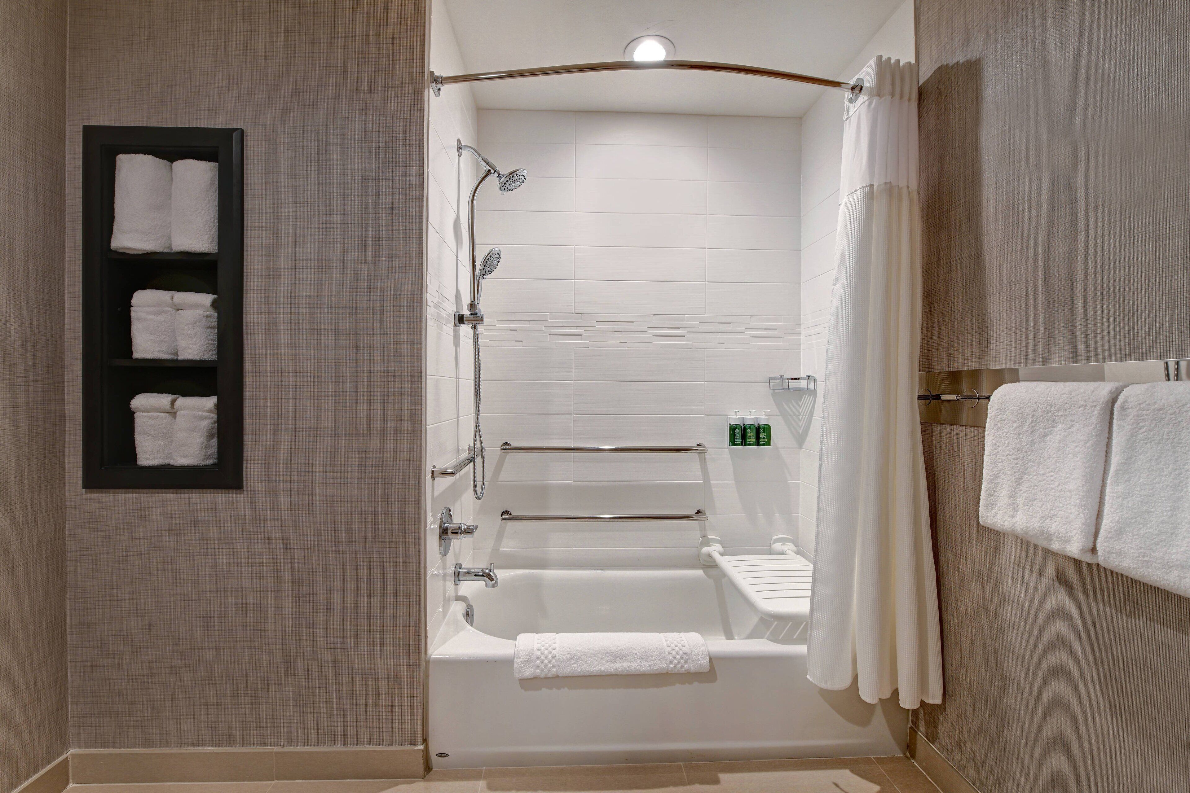 A modern bathroom with a bathtub, shower, and toilet. There are towels on the wall and a shelf with toiletries.