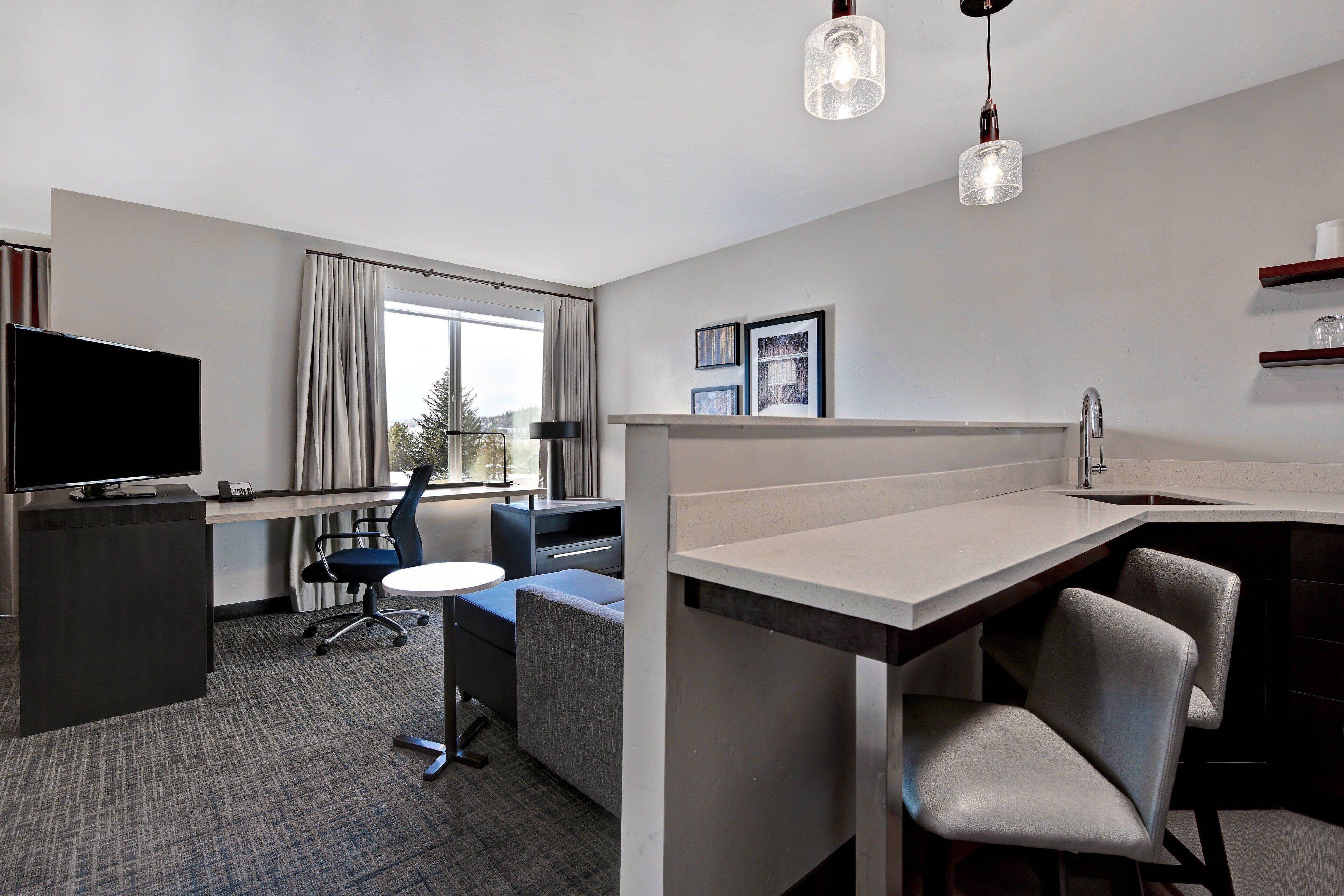 A modern hotel room with a flat-screen TV, a desk with a chair, a small round table, and a kitchenette area.