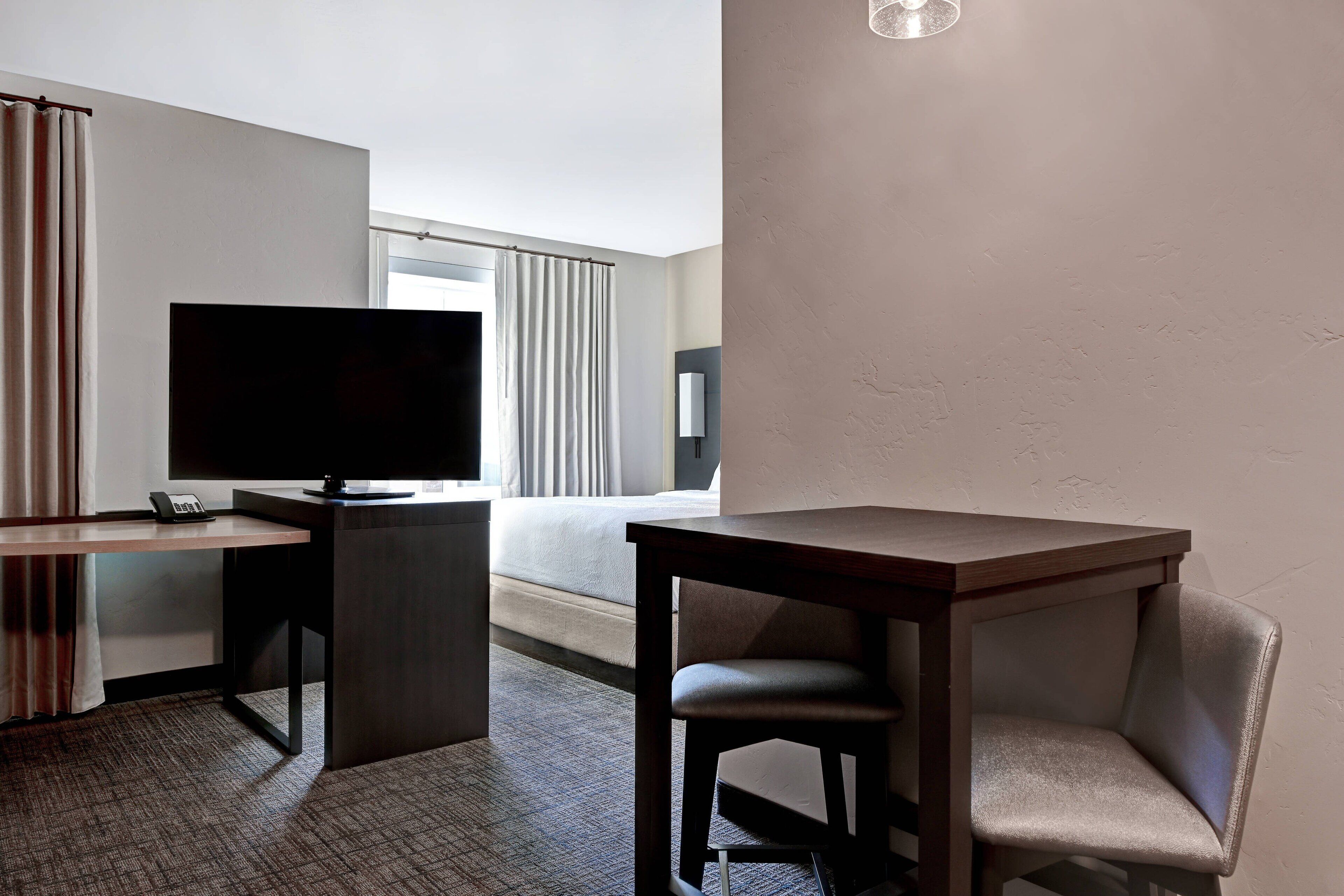 A hotel room with a bed, a desk with a flat-screen TV, a chair, and a small table.