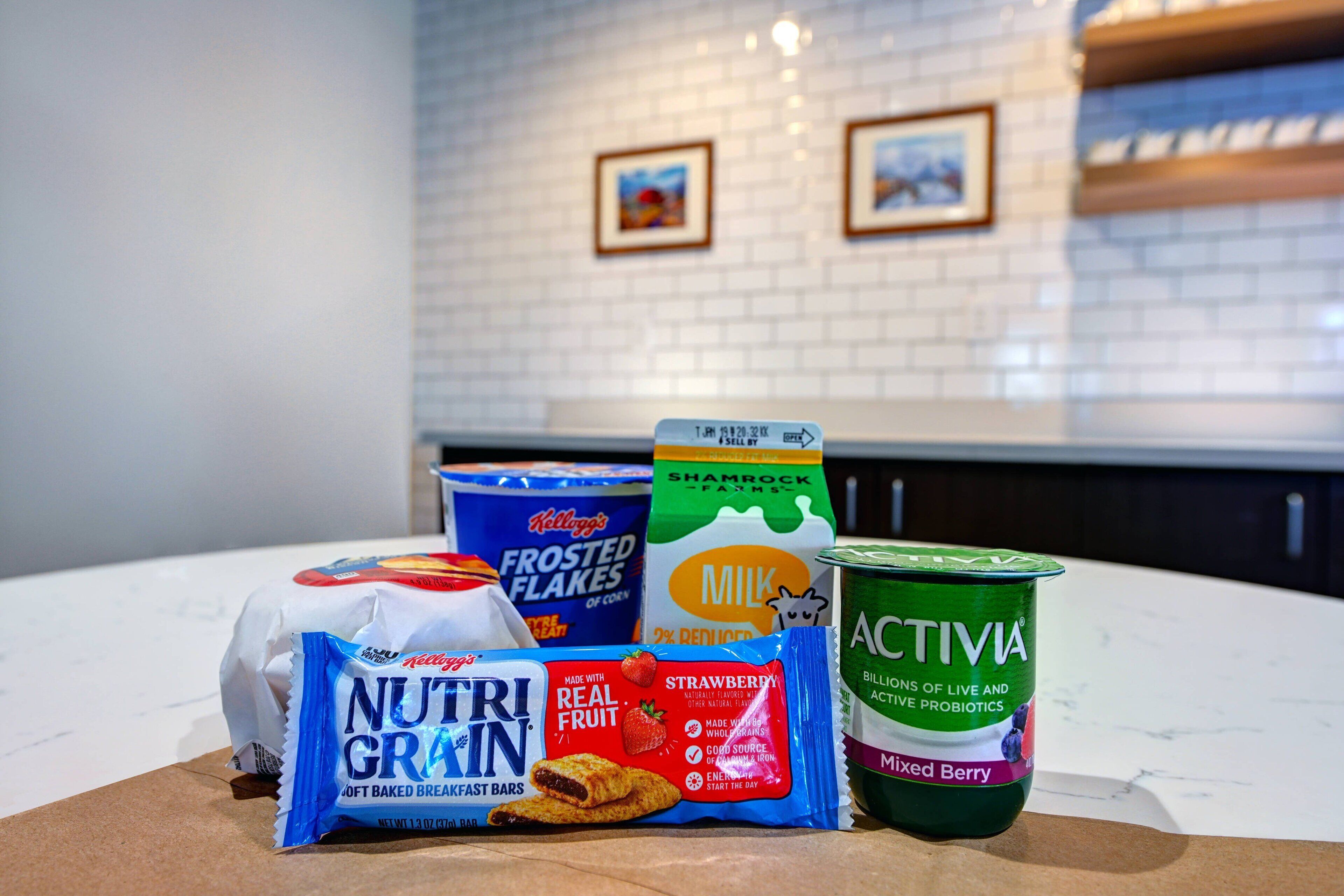 A kitchen counter with various food items including a bag of Nutri-Grain breakfast bars, a carton of milk, a container of frozen flakes, and a container of Activia yogurt.