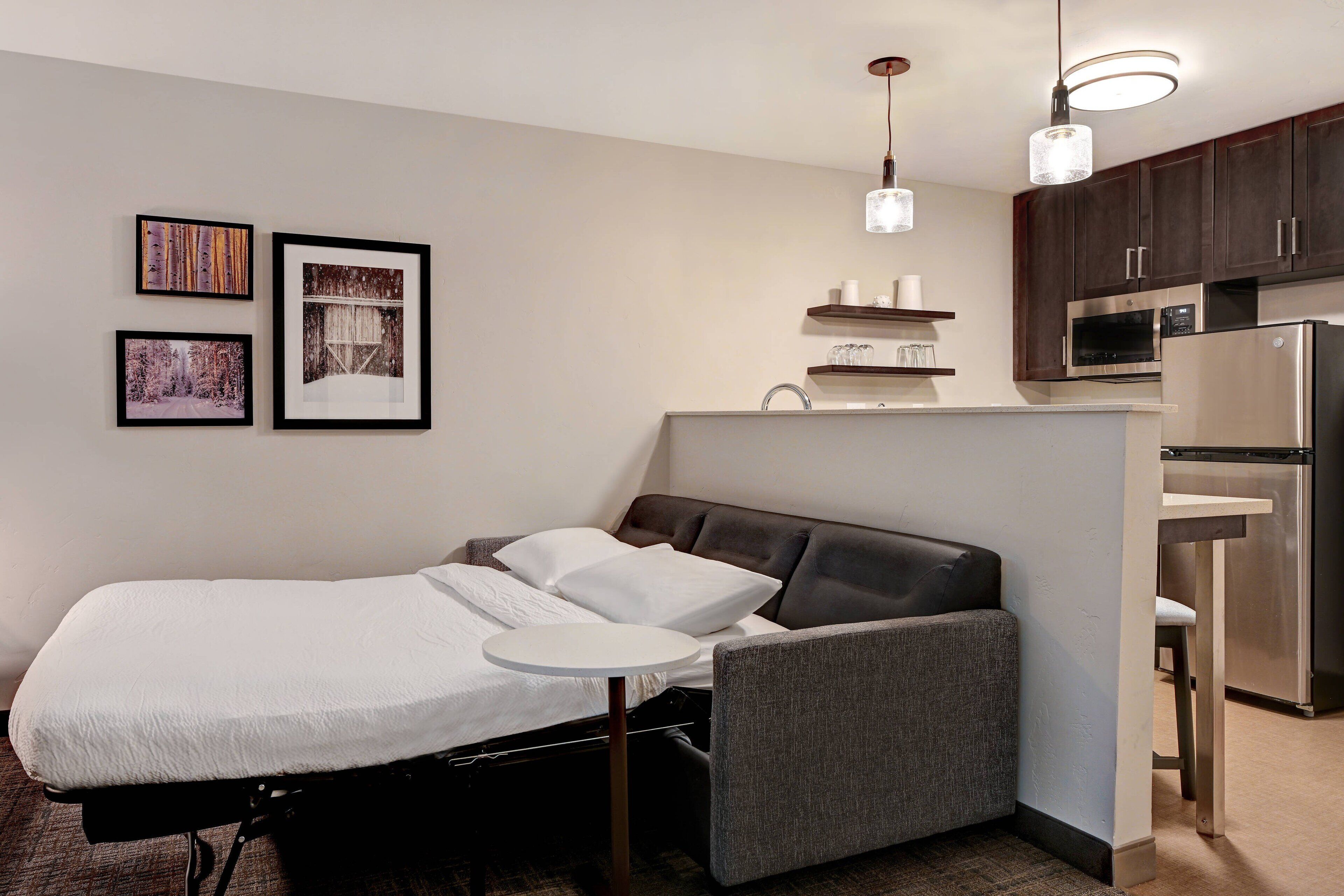 A compact room with a bed, a small table, a kitchenette, and framed pictures on the wall.
