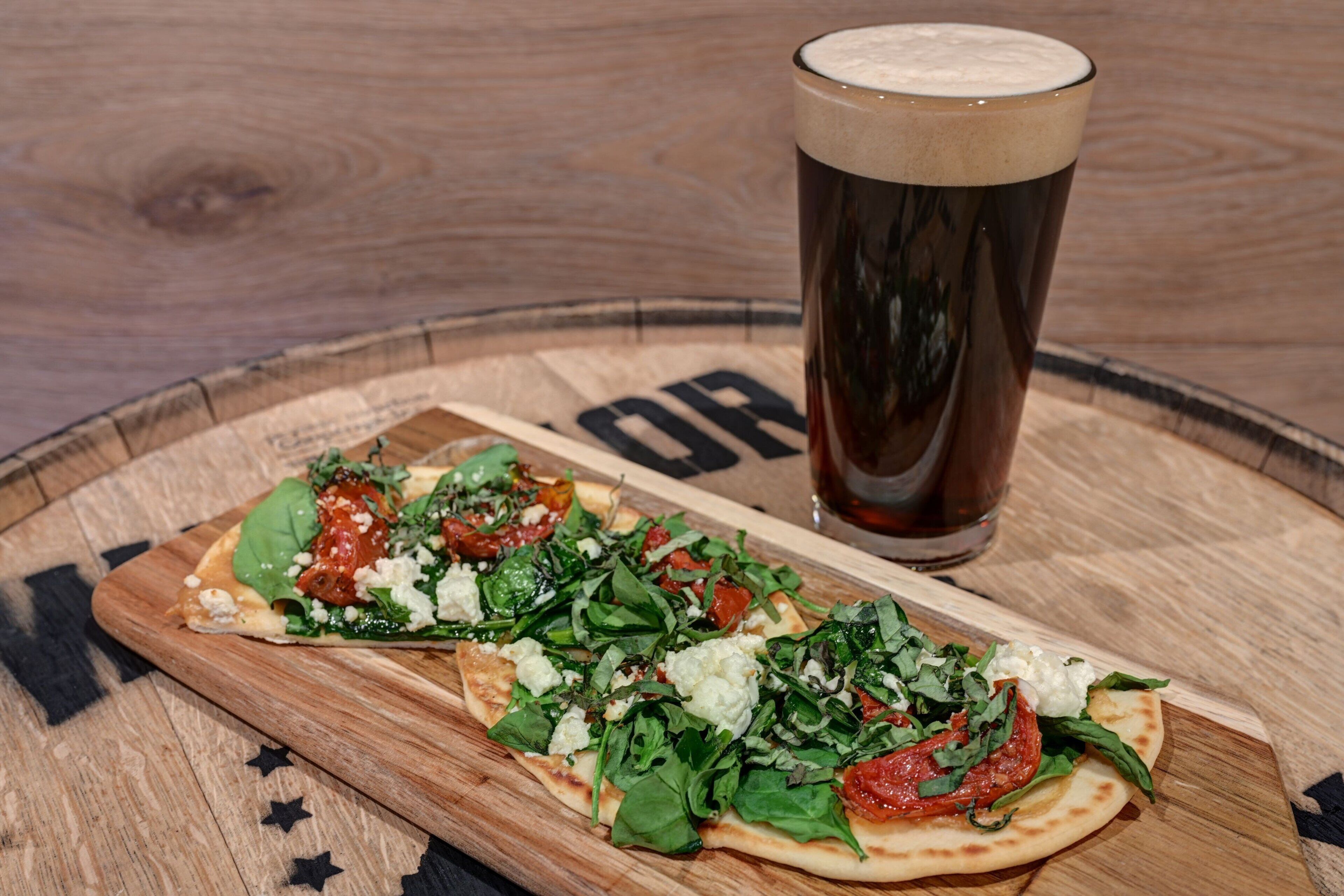 Two pizzas on a wooden cutting board with a glass of beer beside them.