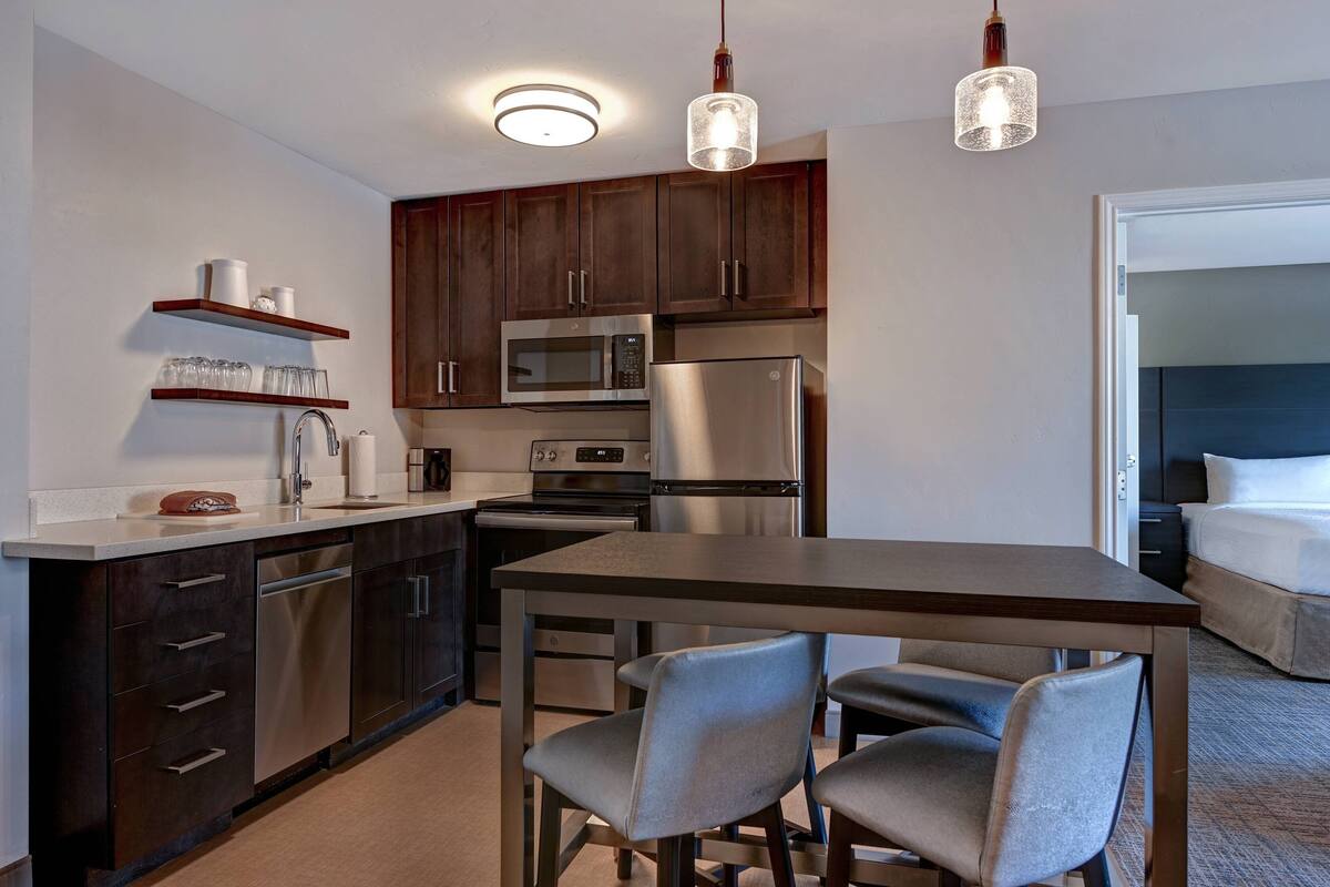 A compact kitchen with modern appliances, a dining area with two chairs, and a bed in the adjacent room.