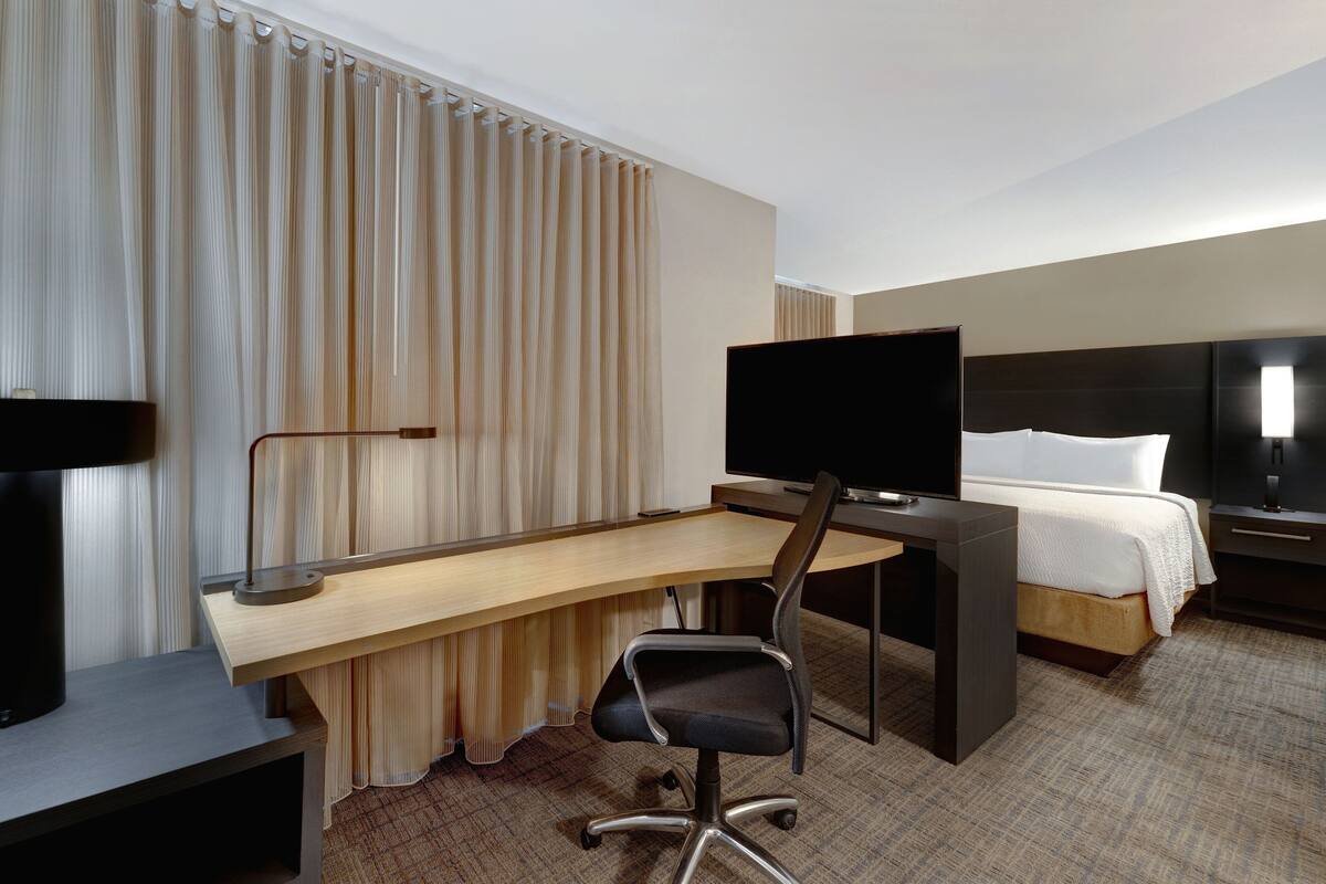 A hotel room with a desk, chair, television, bed, and a large window with curtains.