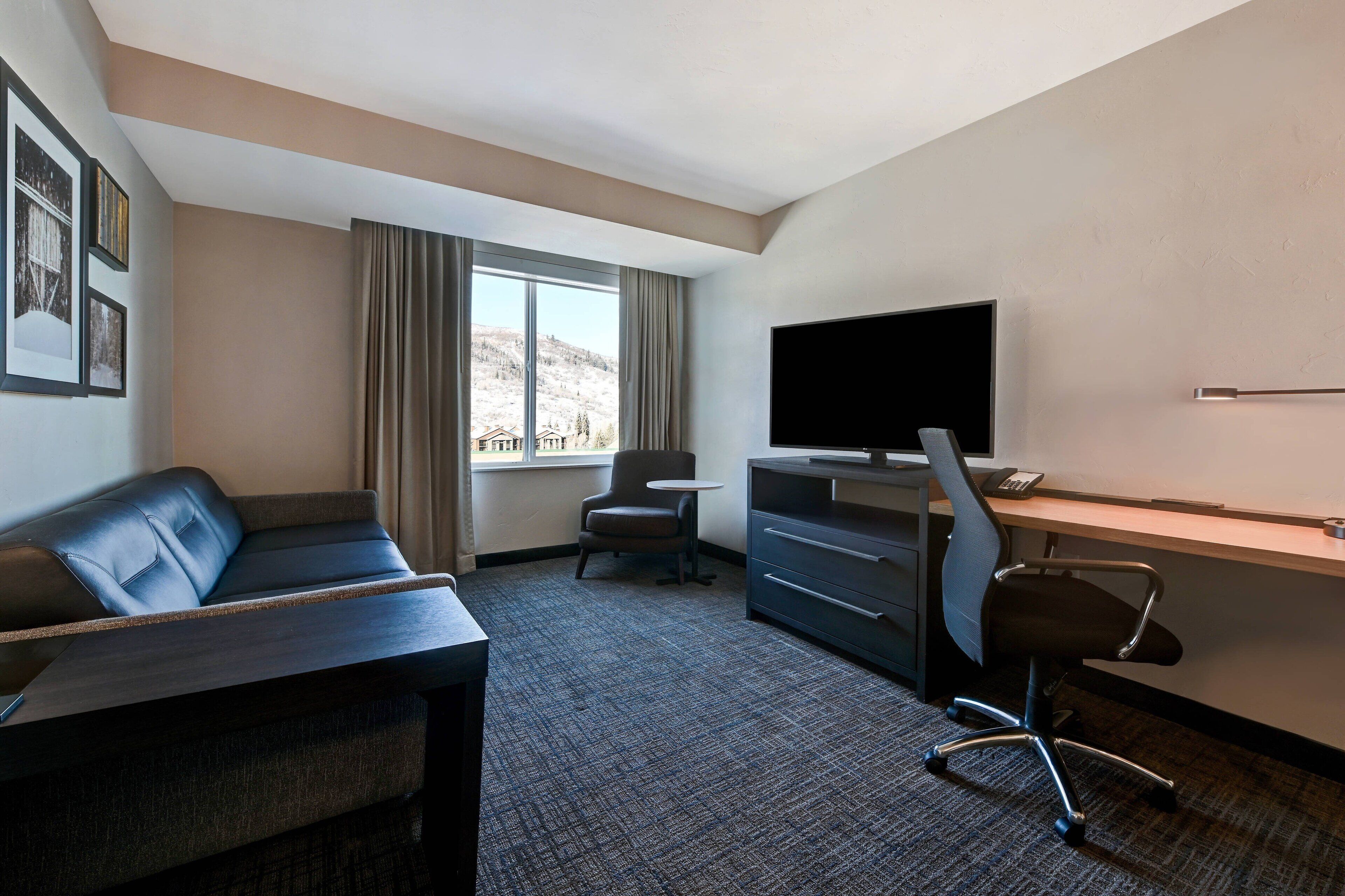 Ett hotellrum med en soffa, ett skrivbord, en stol, en tv och ett fönster med gardiner.