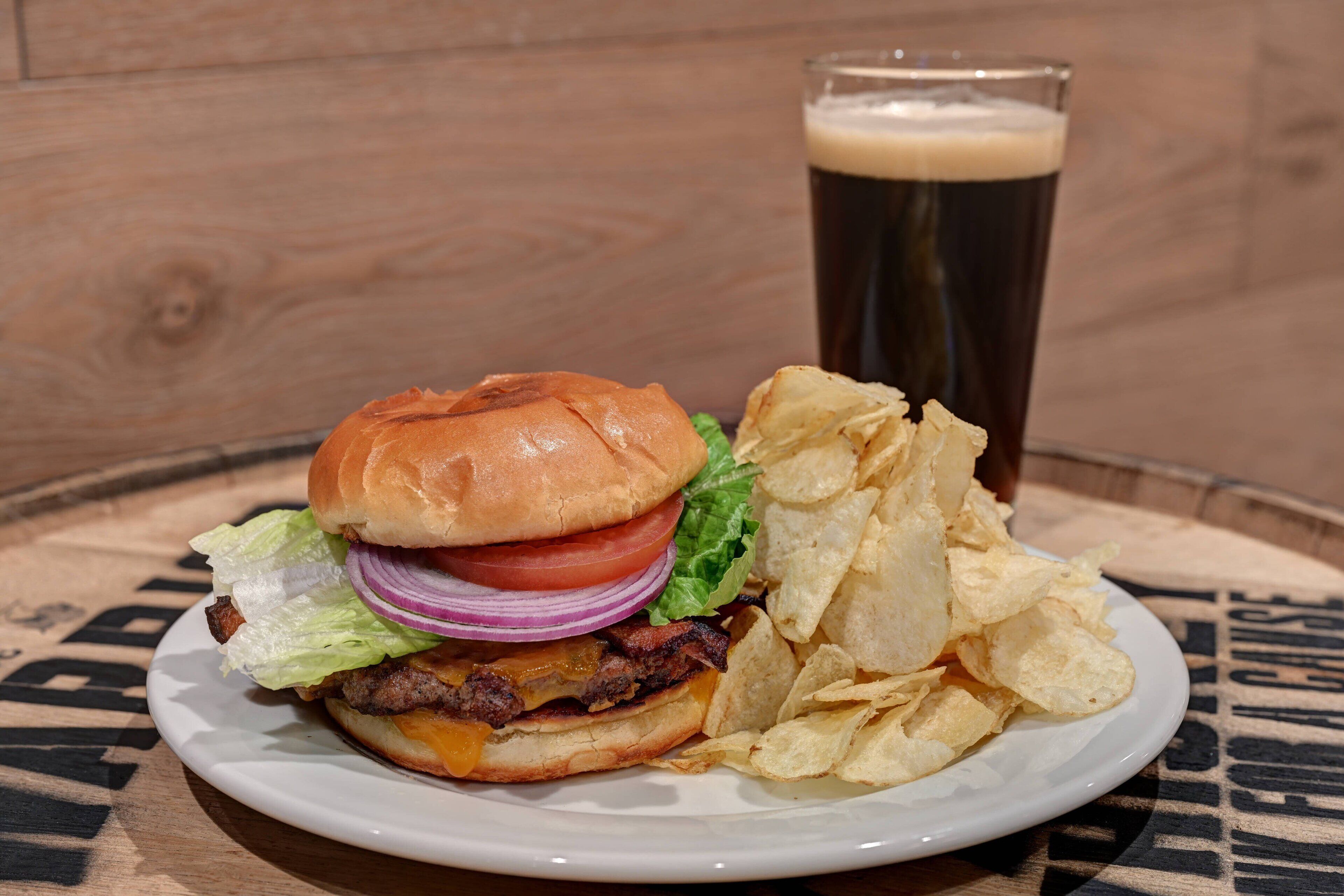 A plate with a burger, chips, and a glass of dark beer on a wooden table.
