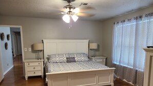 A bedroom with a wooden floor, a white bed with patterned bedding, two bedside tables with lamps, a ceiling fan, and a window with sheer curtains. - Beautiful Lake Front home w/ sunroom and outdoor patio, (Stigler)