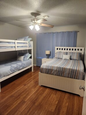 A bedroom with two bunk beds, a single bed, a ceiling fan, a nightstand with a lamp, and a window with curtains.