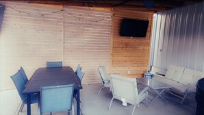 A covered outdoor patio with a wooden table and chairs, a sofa, and a flat-screen TV mounted on the wall. - Beautiful Lake Front home w/ sunroom and outdoor patio, (Stigler)
