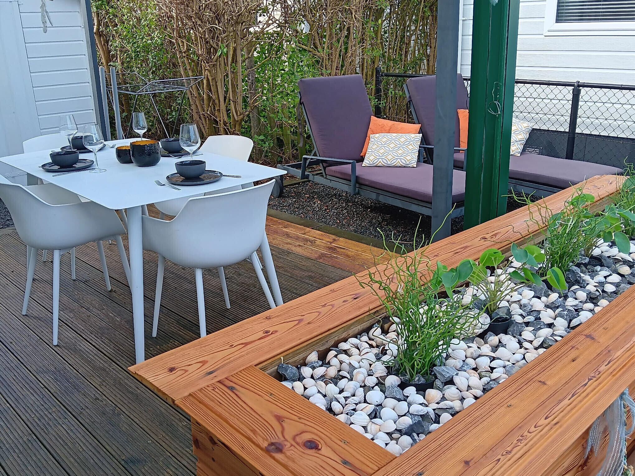 A wooden deck with a white table and chairs, surrounded by a garden bed with plants and pebbles.