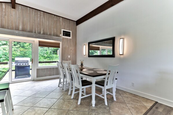 A dining area with a table and chairs, a large window, and a view of greenery outside. - Breakaway House: Killington Duplex, Unit A. Hot Tub Close to Resort. (Killington)