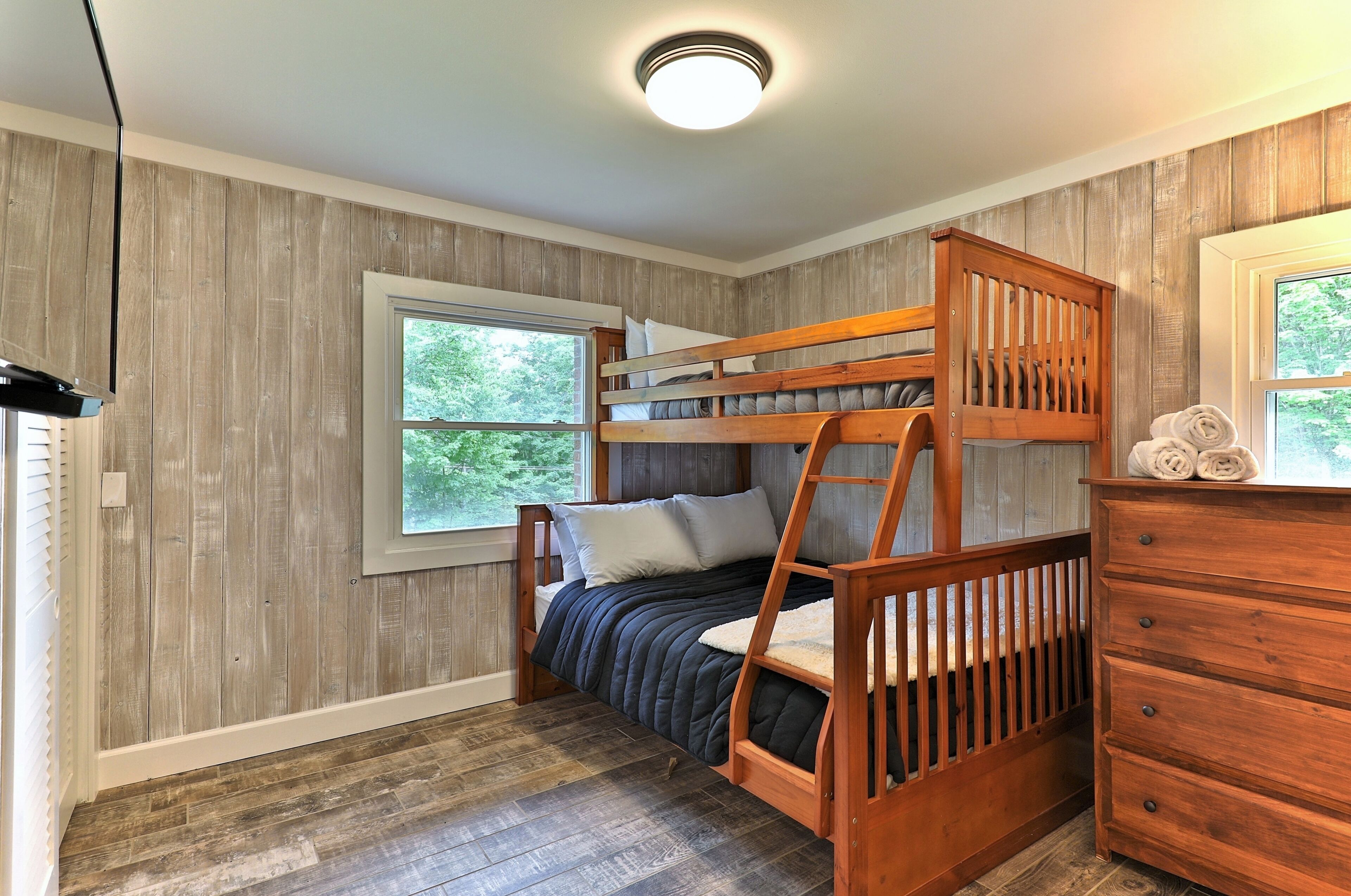 A bunk bed with a wooden ladder, a dresser, and a window with a view of trees.