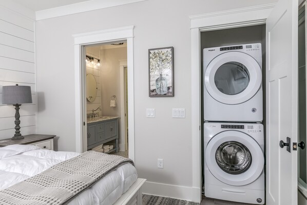 A bedroom with a bed, a lamp, a washing machine, and a bathroom visible through an open door.