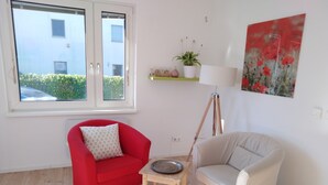 A room with a red armchair, a beige armchair, a small wooden table, a floor lamp, and a window with a view of buildings and greenery.