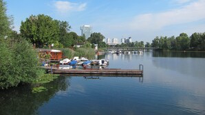 A calm lake with boats docked at a pier, surrounded by greenery and a city skyline in the background.