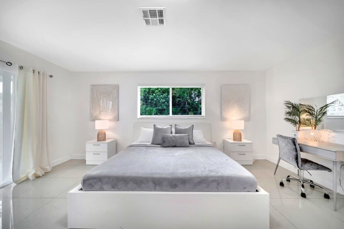 A modern bedroom with a large bed, bedside tables with lamps, a desk, and a window with a view of greenery.