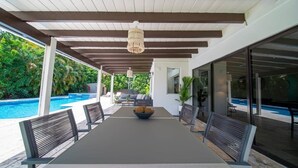 Un espace extérieur abrité, aménagé pour les repas, avec une table et des chaises, un luminaire suspendu, et une vue sur une piscine et la verdure.
