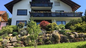 A two-story building with a balcony on the upper floor, surrounded by greenery and rocks.