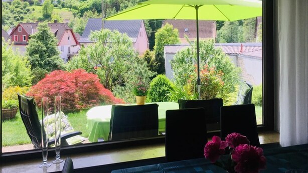 A room with a large window overlooking a garden and houses. There is a table with chairs and a green umbrella.