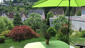 A patio with a green table and umbrella, surrounded by lush greenery and houses in the background.