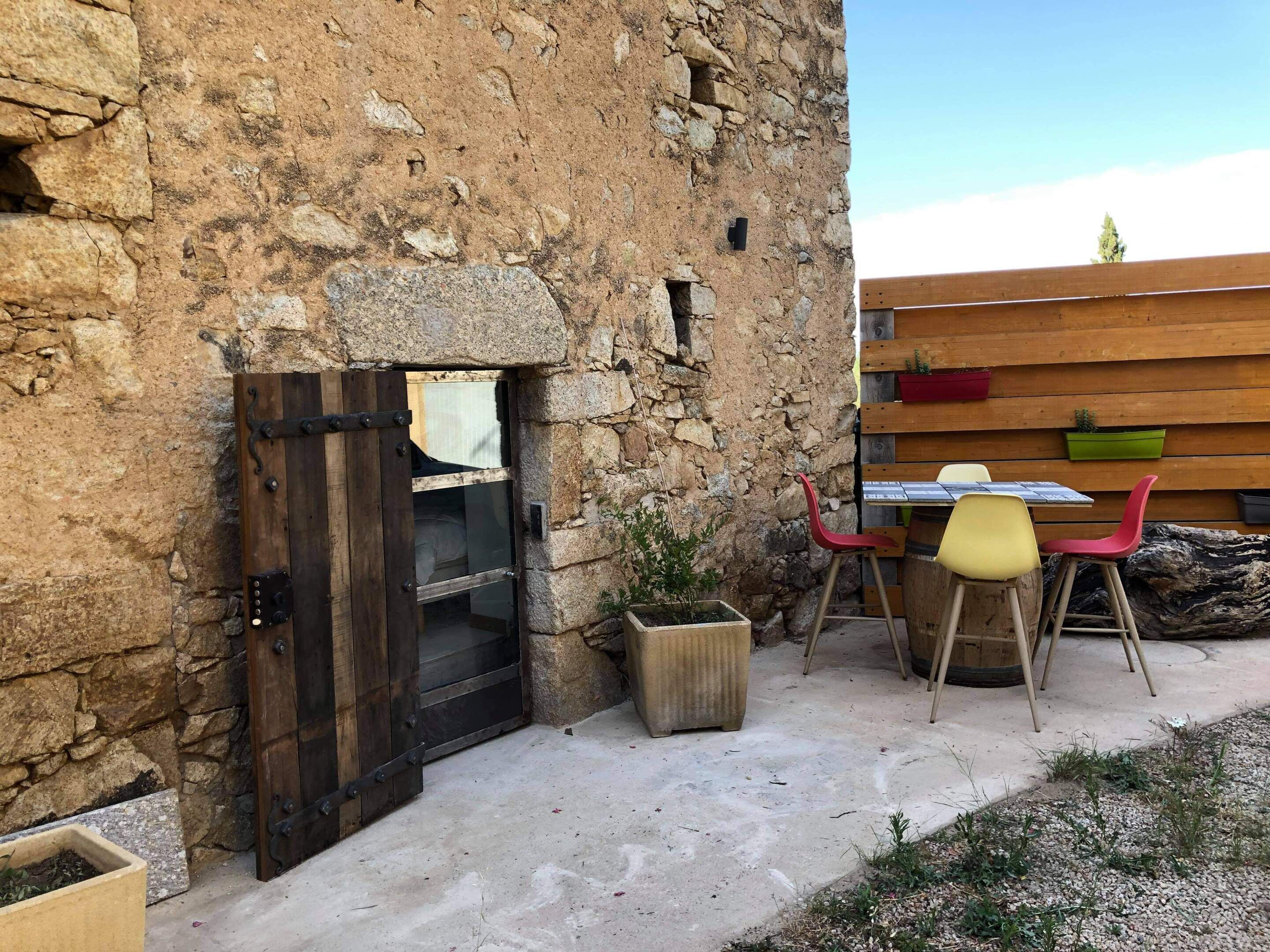 A rustic stone building with a wooden door, a table with chairs, and potted plants.