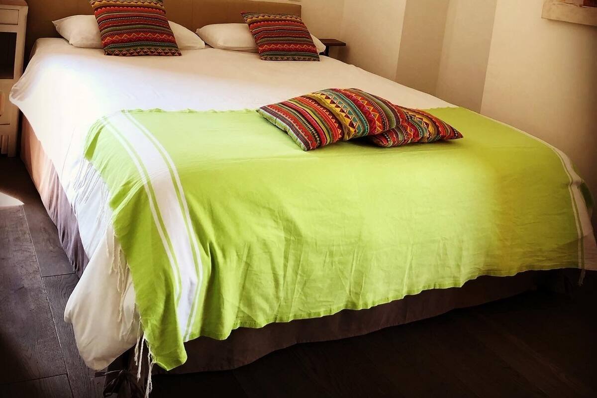 Una cama bien tendida con una colcha verde y dos almohadones decorativos, una mesita de noche con una lámpara, y un sencillo arco de entrada.