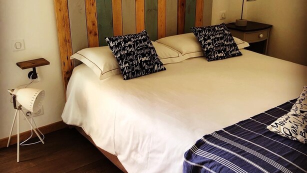 A bed with white bedding, two black and white patterned pillows, a wooden headboard, a bedside lamp, and a small bedside table with a lamp. - 1722 Chambres de Charme (Ville-di-Paraso)