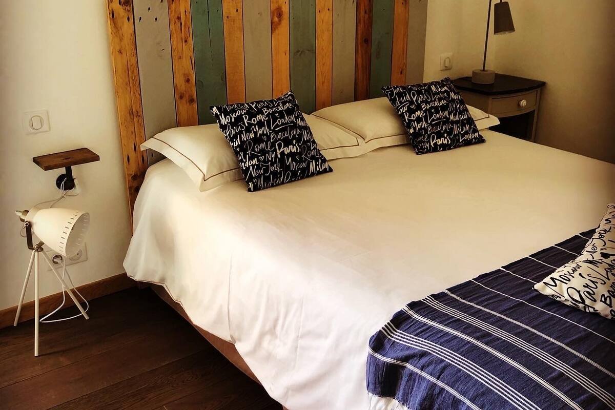 Una cama con ropa blanca, dos almohadas con estampado blanco y negro, un cabecero de madera, una lámpara de noche y una mesita de noche con una lámpara.