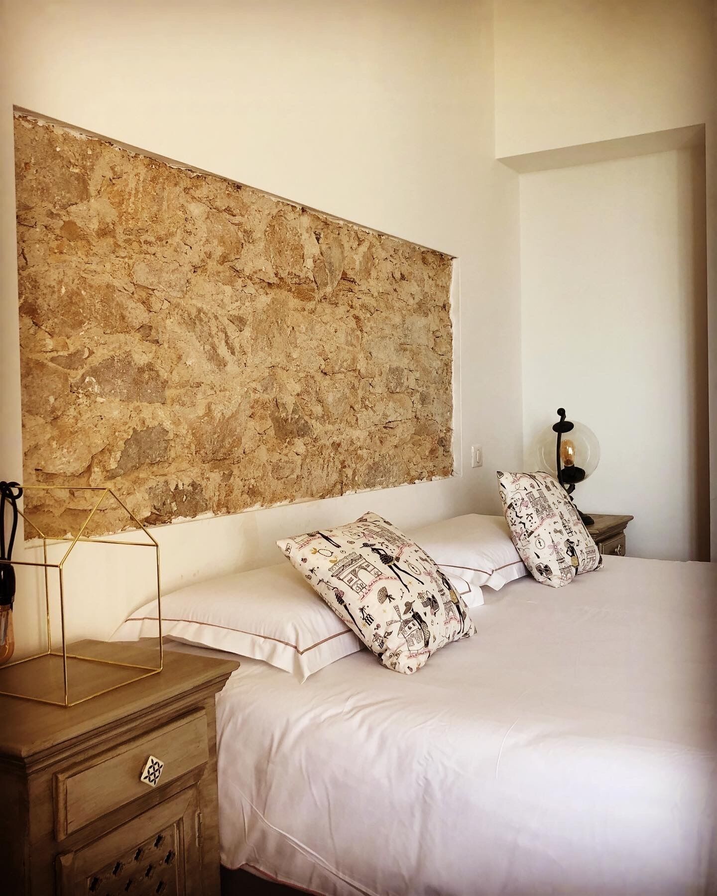A bedroom with a large stone wall, a wooden nightstand, a lamp, and patterned pillows.