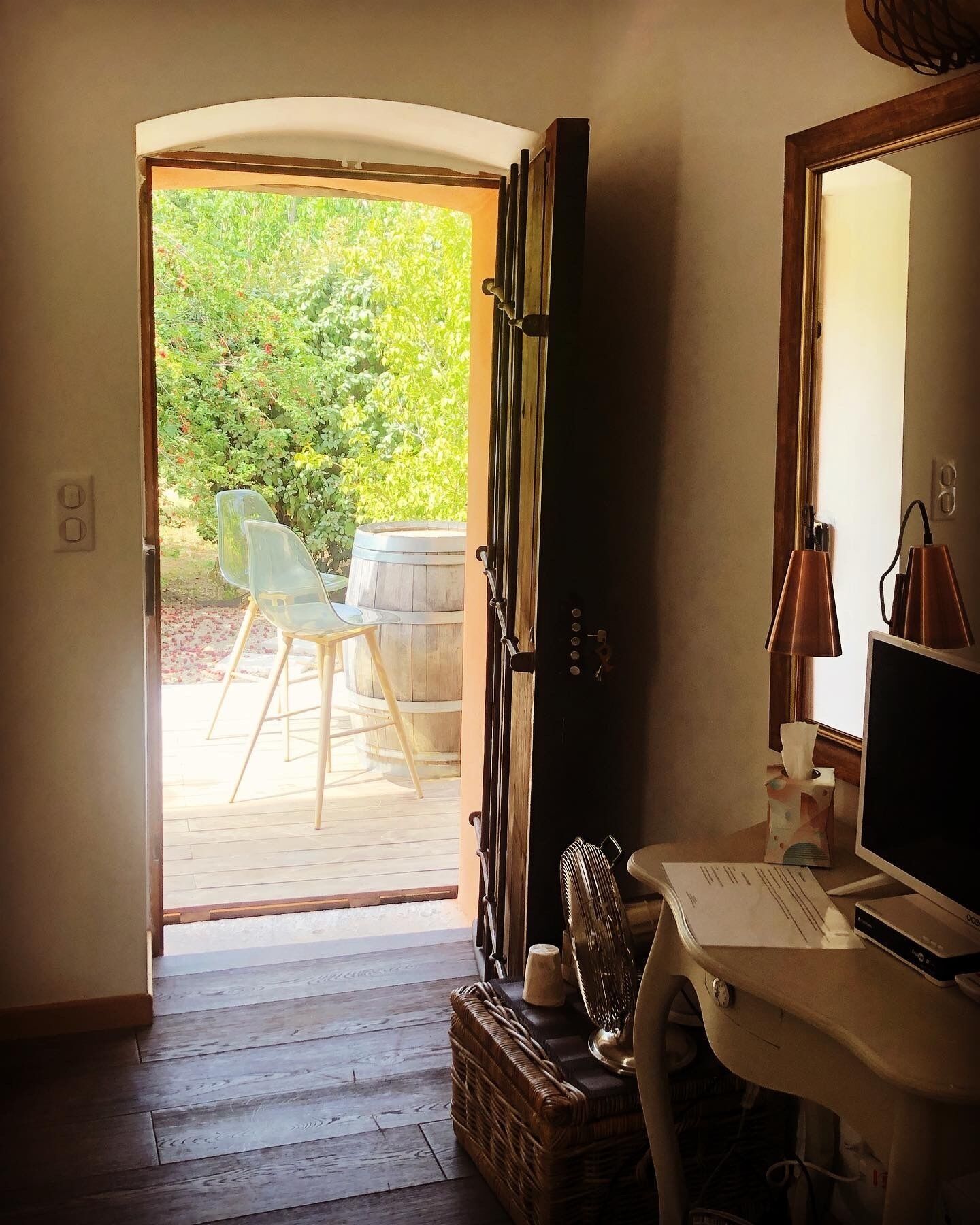 Una stanza con una scrivania in legno, un computer, una sedia e una finestra che si apre su un'area esterna con sedie e una botte di legno.