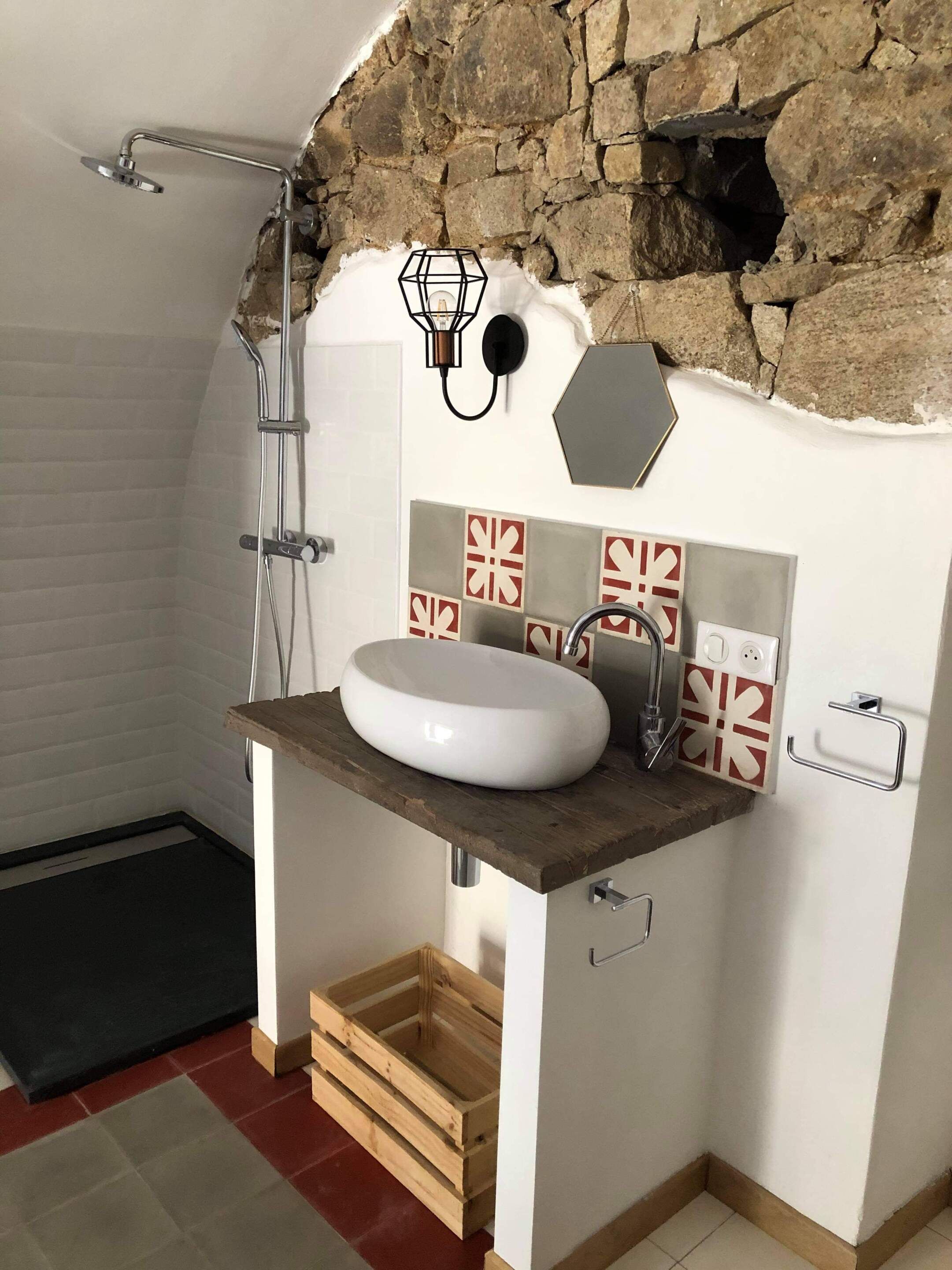 Un baño con pared de piedra, tocador de madera, lavamanos blanco, ducha y un pequeño cofre de madera.