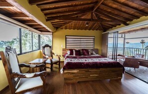 Ein Schlafzimmer mit einem hölzernen Bett, einem Sessel, einem Tisch und großen Fenstern.