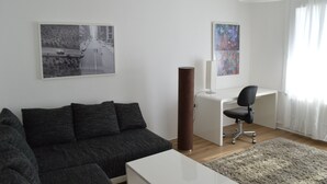 Un saló modern amb un sofà gris fosc, una taula de centre blanca, un escriptori amb una cadira i un quadre abstracte gran a la paret.