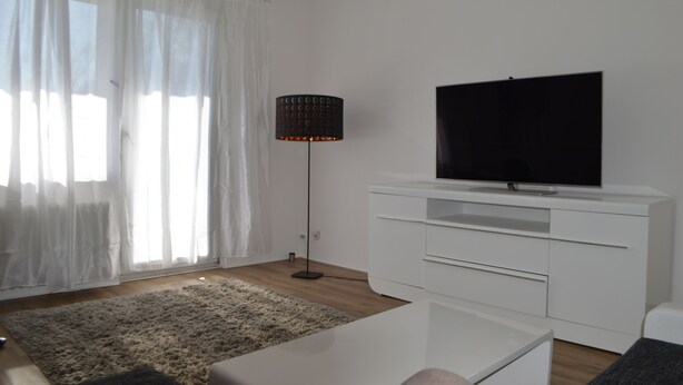 Un saló modern amb televisió de pantalla plana, mobles blancs, una làmpada de peu i cortines transparents.
