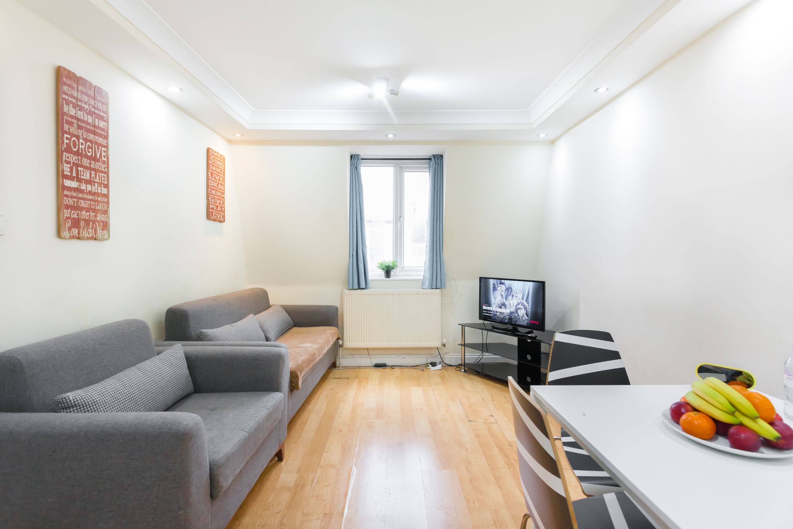 Una sala de estar moderna con un sofá gris, una mesa de comedor con fruta, un televisor y un ventanal con cortinas.
