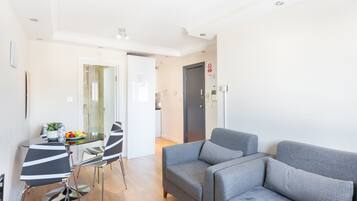 A modern living room with a grey sofa, a glass dining table with striped chairs, and a kitchen in the background.