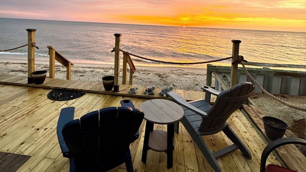 Une terrasse en bois, agrémentée de chaises et de tables, d’où l’on peut admirer l’océan au coucher du soleil.