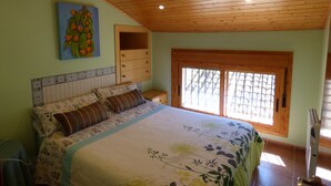 A bedroom with a wooden ceiling, a bed with floral bedding, a nightstand, a window with a view, and a wall-mounted shelf.