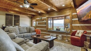 A cozy log cabin interior with a sectional sofa, a red armchair, a wooden coffee table, and a large window offering a mountain view.