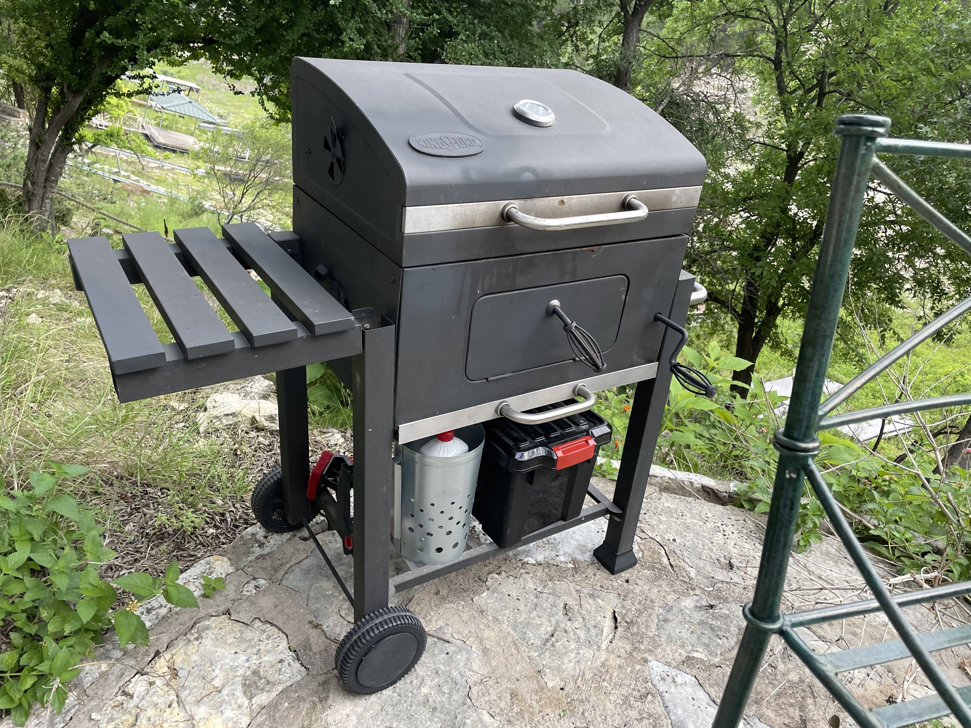 Ein tragbarer Grill mit Rädern und Stauraum, auf einer Steinoberfläche mit Grünpflanzen im Hintergrund.