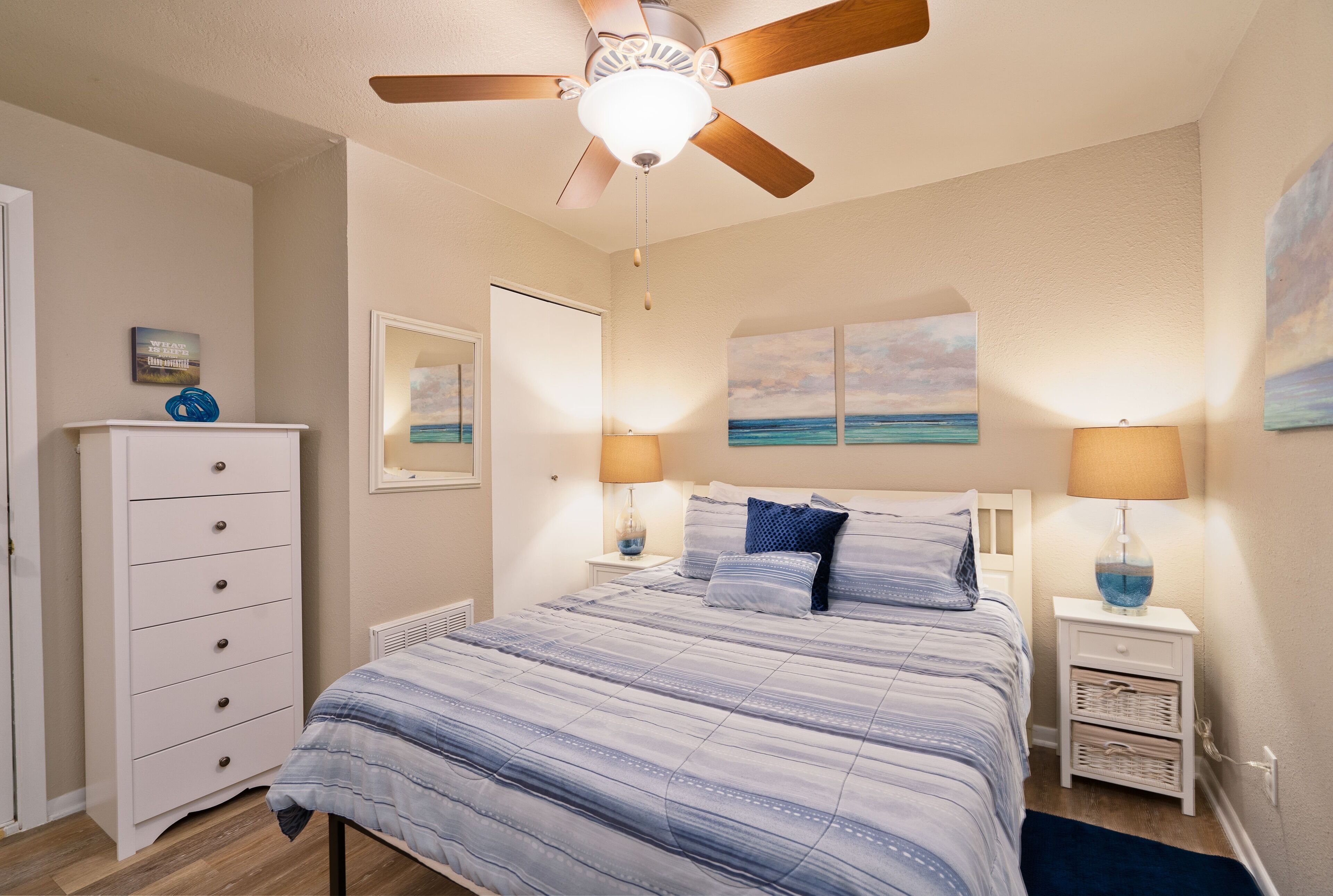 A bedroom with a bed, a ceiling fan, a dresser, and artwork on the walls.