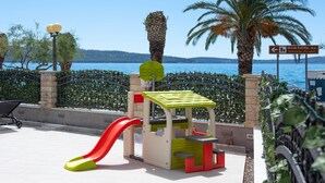 A playground with a slide and climbing structures, surrounded by a fence and palm trees, with a view of the sea and a sign indicating 'Utrda Kastilac, 16th century'. - Beach front apartment in Kastel Gomilica (Kastel Gomilica)