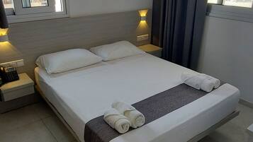 Un letto rifatto con una coperta grigia e asciugamani arrotolati, un comodino con una lampada e una finestra con tende.