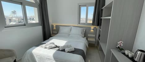 A modern hotel room with a large bed, two bedside tables, a desk with a kettle, and a view of the cityscape through the windows.