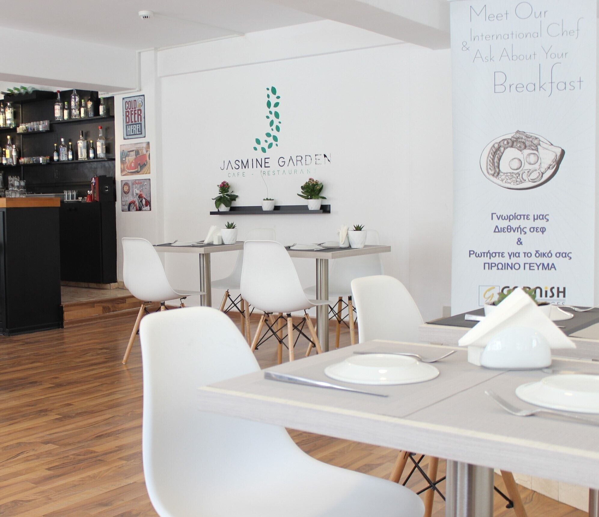 A modern cafe interior with white furniture, a bar area with liquor bottles, and a wall sign for 'Jasmine Garden Cafe - Restaurant'.