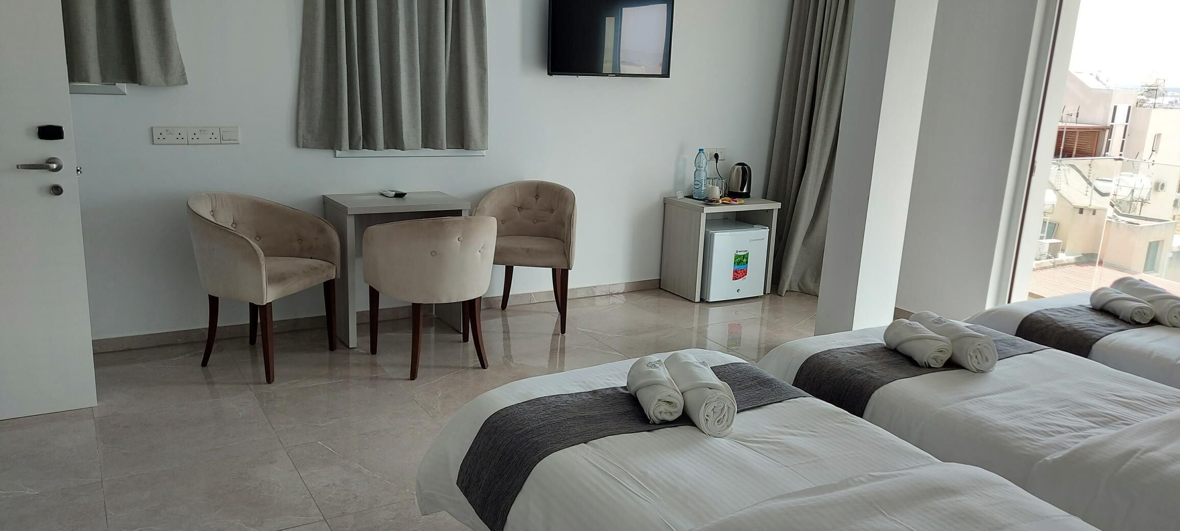 Ein Hotelzimmer mit zwei Betten, einem Schreibtisch, zwei Stühlen, einer Minibar und einem Wandfernseher.